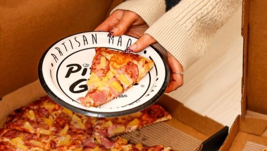 This photo shows a woman's hands holding a plate with a slice of Hawaiian pizza.