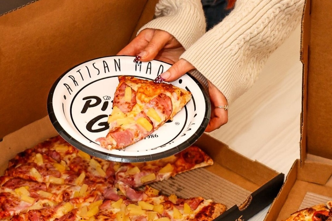 This photo shows a woman's hands holding a plate with a slice of Hawaiian pizza.