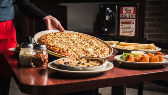 This photo shows a large pizza being placed on a table at a Pizza King location. There is also a black phone for table side ordering with text that reads, Ring the King.