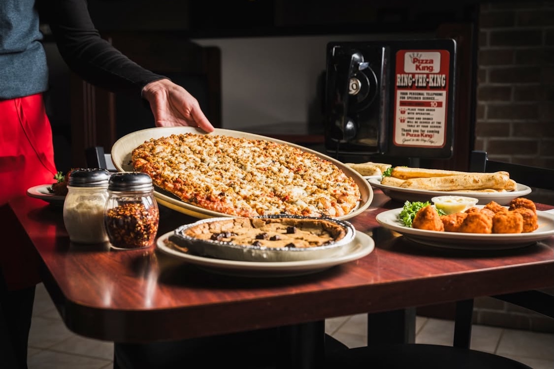 This photo shows a large pizza being placed on a table at a Pizza King location. There is also a black phone for table side ordering with text that reads, Ring the King.