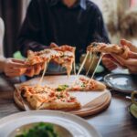 This photo shows a group of pizzeria customers, shown from the neck down, removing slices of pizza from whole pies.