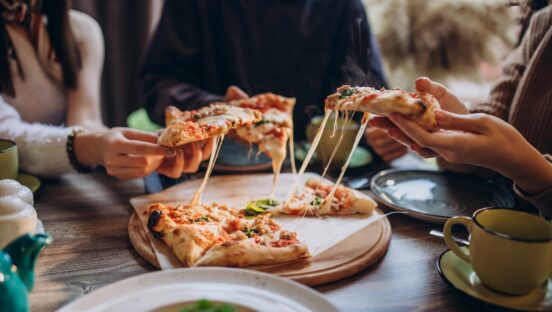 This photo shows a group of pizzeria customers, shown from the neck down, removing slices of pizza from whole pies.