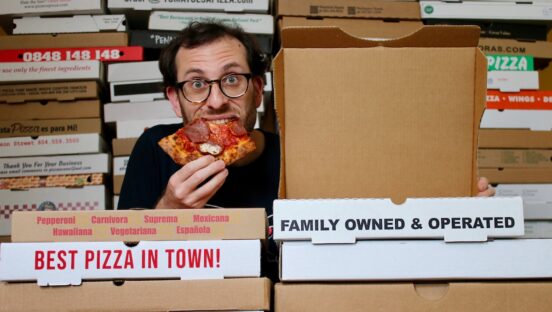 This photo shows Scott Wiener crouched amongst a huge stack of pizza boxes, biting into a slice, with one box open next to him.