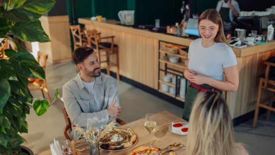 This photo shows a smiling waitress serving gourmet pizza to happy customers in a pizzeria.