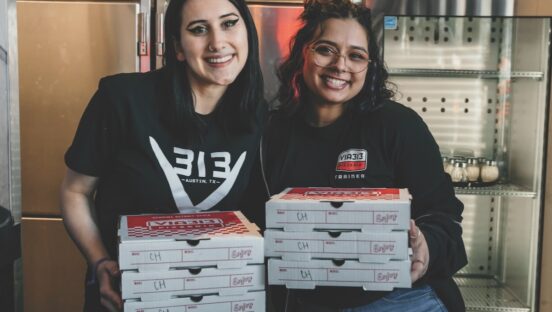This photo shows two smiling women in Via 313 shirts holding pizza boxes in their hands.