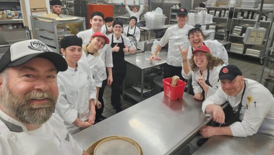 This photo shows Will Grant, in a white chef coat, in the forefront and to the left, shooting a selfie with a group of happy-looking students.