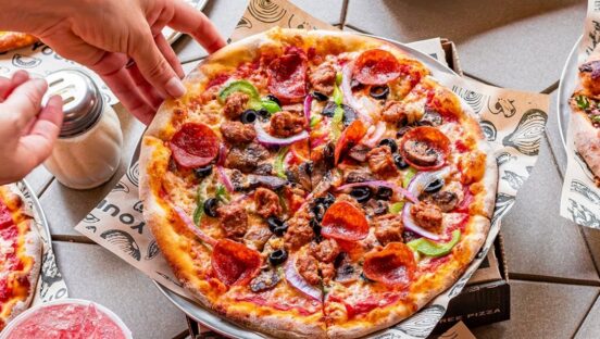 This photo shows a woman's hand reaching for a slice of a whole pizza topped with pepperoni, sausage, black olives, bell peppers and red onions.