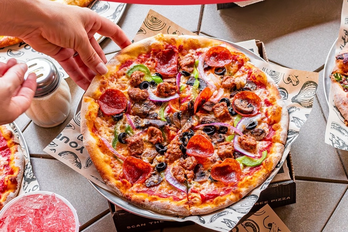 This photo shows a woman's hand reaching for a slice of a whole pizza topped with pepperoni, sausage, black olives, bell peppers and red onions.