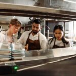 This photo shows a man standing between two women, in a restaurant kitchen, busy with some kind of task that's obscured by a shelf.