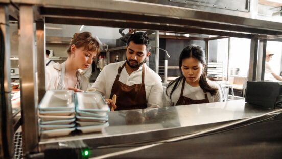 This photo shows a man standing between two women, in a restaurant kitchen, busy with some kind of task that's obscured by a shelf.