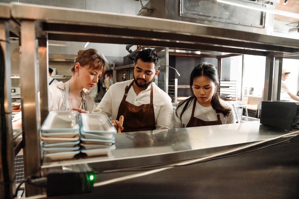 This photo shows a man standing between two women, in a restaurant kitchen, busy with some kind of task that's obscured by a shelf.