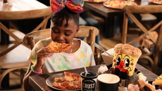 This photo shows a young girl with a red bow in her hair biting into a slice of pizza with beverage cups with the Crust logo on them in front of her.