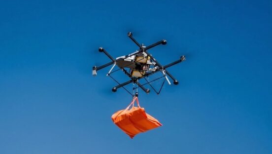 This photo shows a drone, hovering beneath a dark blue sky, carrying an orange bag containing pizzas.