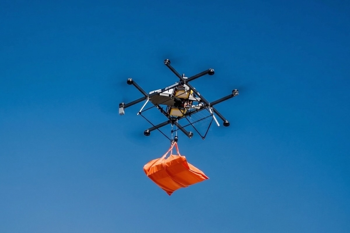 This photo shows a drone, hovering beneath a dark blue sky, carrying an orange bag containing pizzas.