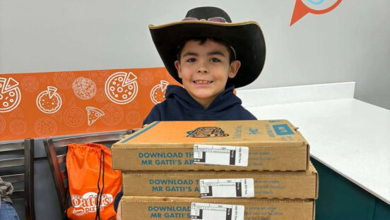This photo shows a little boy wearing a cowboy hat and holding four Mr Gatti's Pizza boxes inside one of the restaurants.