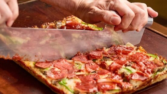 This photo shows a pair of hands slicing a square-shaped Detroit style whole pizza.