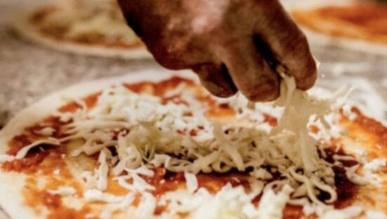 This photo shows a man's hand adding shredded mozzarella to an unbaked pizza skin.