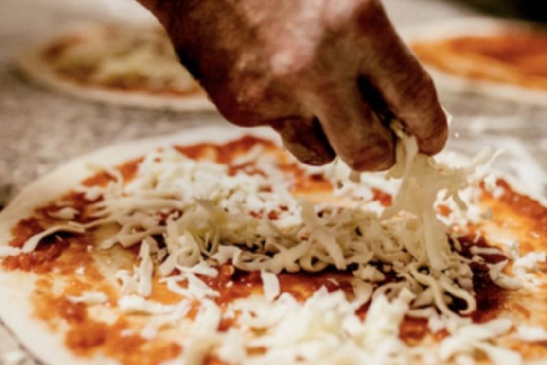 This photo shows a man's hand adding shredded mozzarella to an unbaked pizza skin.