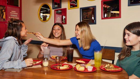 This photo shows four women eating pizza in Pizza Inn. One woman is putting a slice in another woman's mouth, and everyone looks delighted to be there.