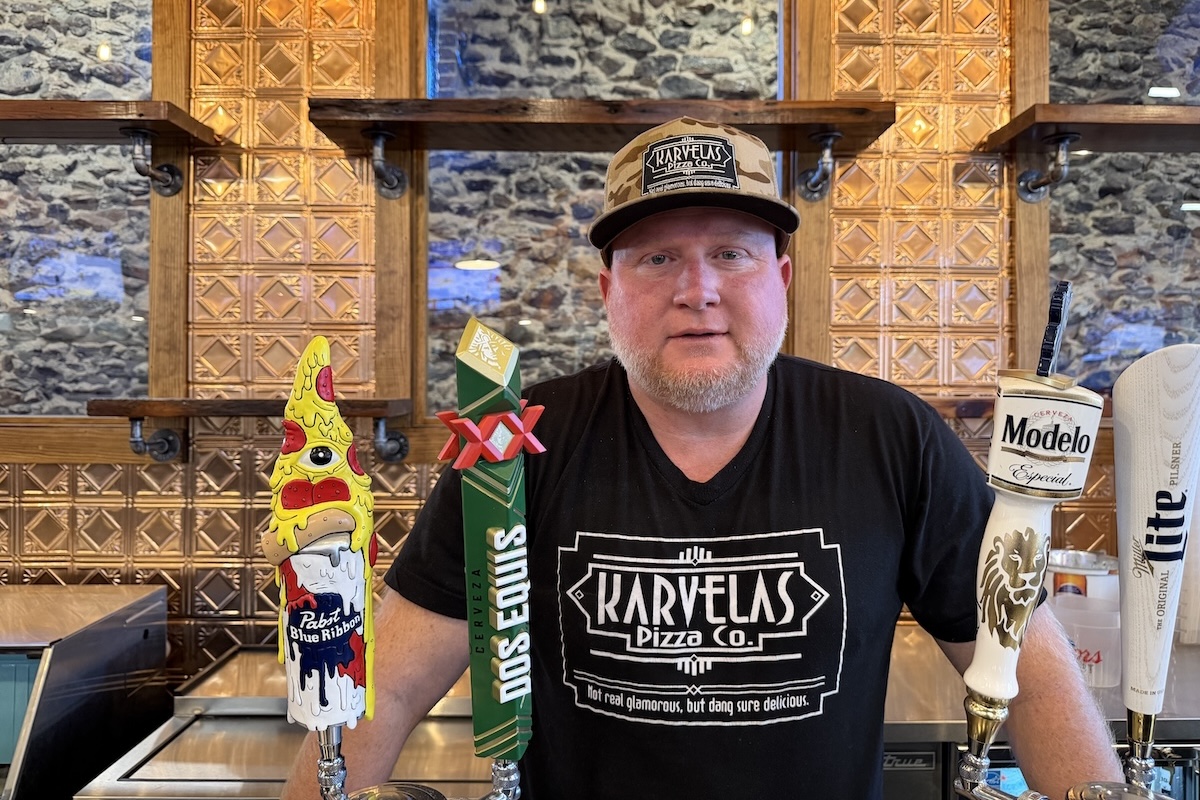 This photo shows Joey Karvelas, in a green camo cap and black Karvelas Pizza Co. t-shirt, standing behind the bar at one of his restaurants.