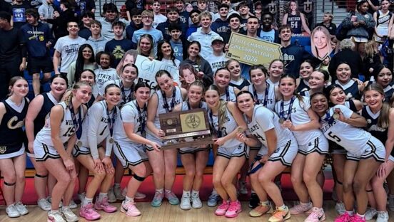 This photo shows a high school girls' basketball team celebrating winning a state championship, surrounded by their fans in the high school gym.