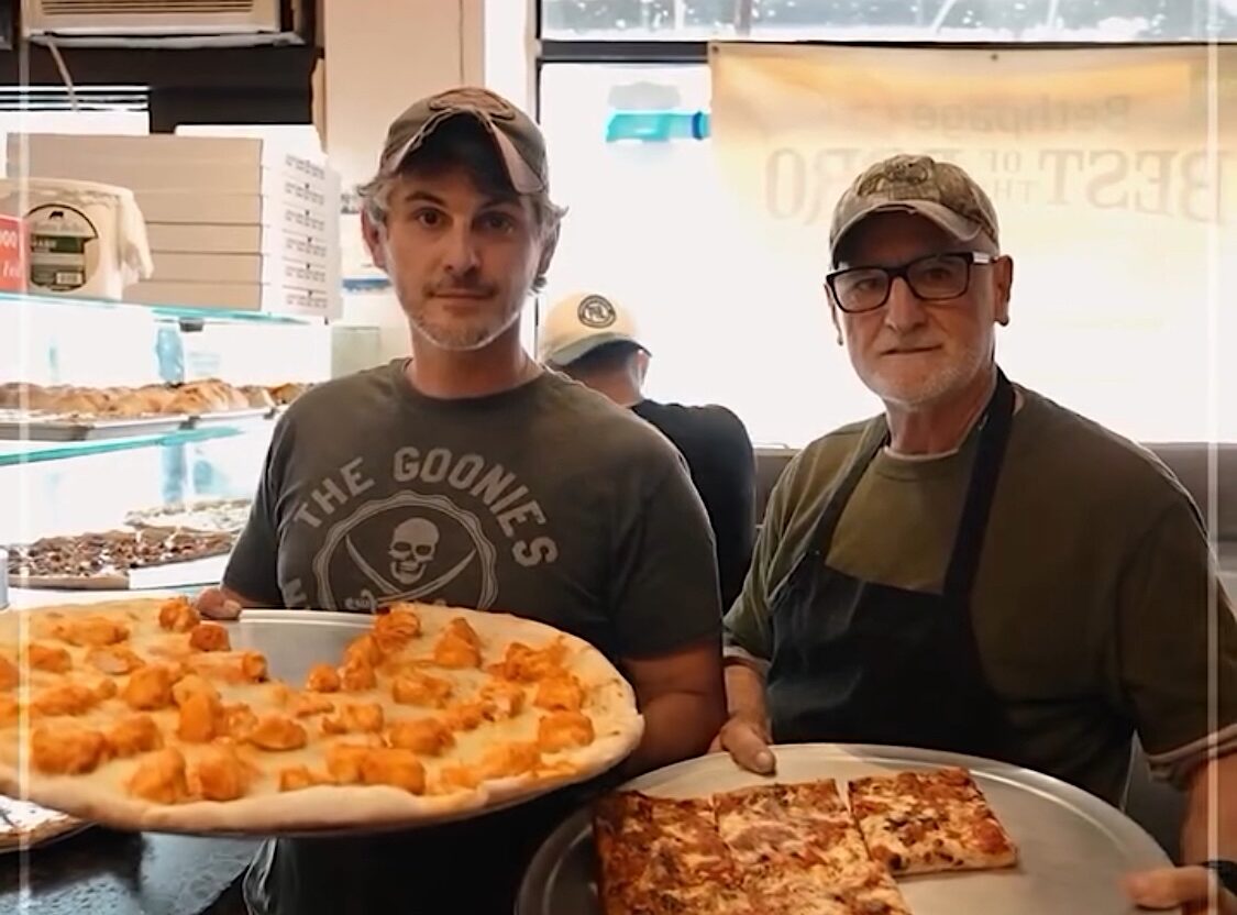 This photo shows Louie and Cazim Suljovic holding large pizzas and looking at the camera in their restaurant, Louie's Pizza.
