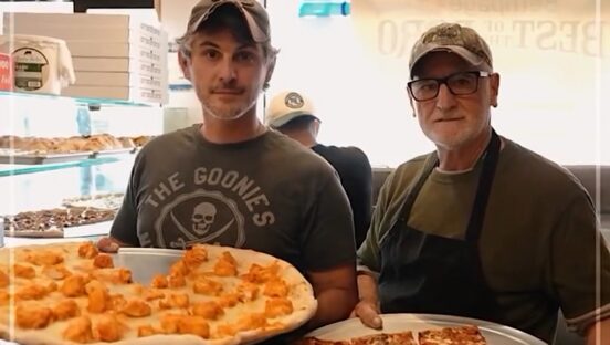 This photo shows Louie and Cazim Suljovic holding large pizzas and looking at the camera in their restaurant, Louie's Pizza.