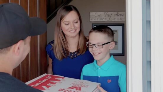 This photo shows a woman and a young boy standing in the doorway of their home, happily receiving a pizza delivery from Master Pizza.