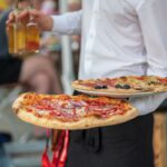 This photo shows a restaurant server, seen from the shoulders down, carrying two pizzas and a glass of beer to a table.