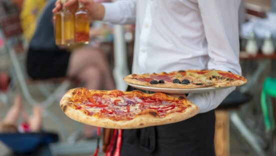 This photo shows a restaurant server, seen from the shoulders down, carrying two pizzas and a glass of beer to a table.