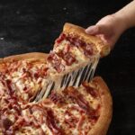 This photo shows a person's hand removing a slice from a triple bacon pizza from Papa Johns.