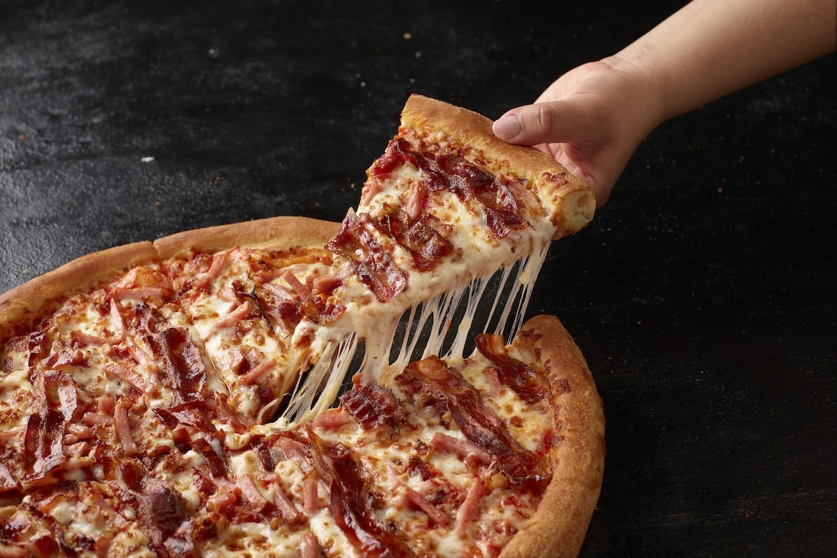This photo shows a person's hand removing a slice from a triple bacon pizza from Papa Johns.