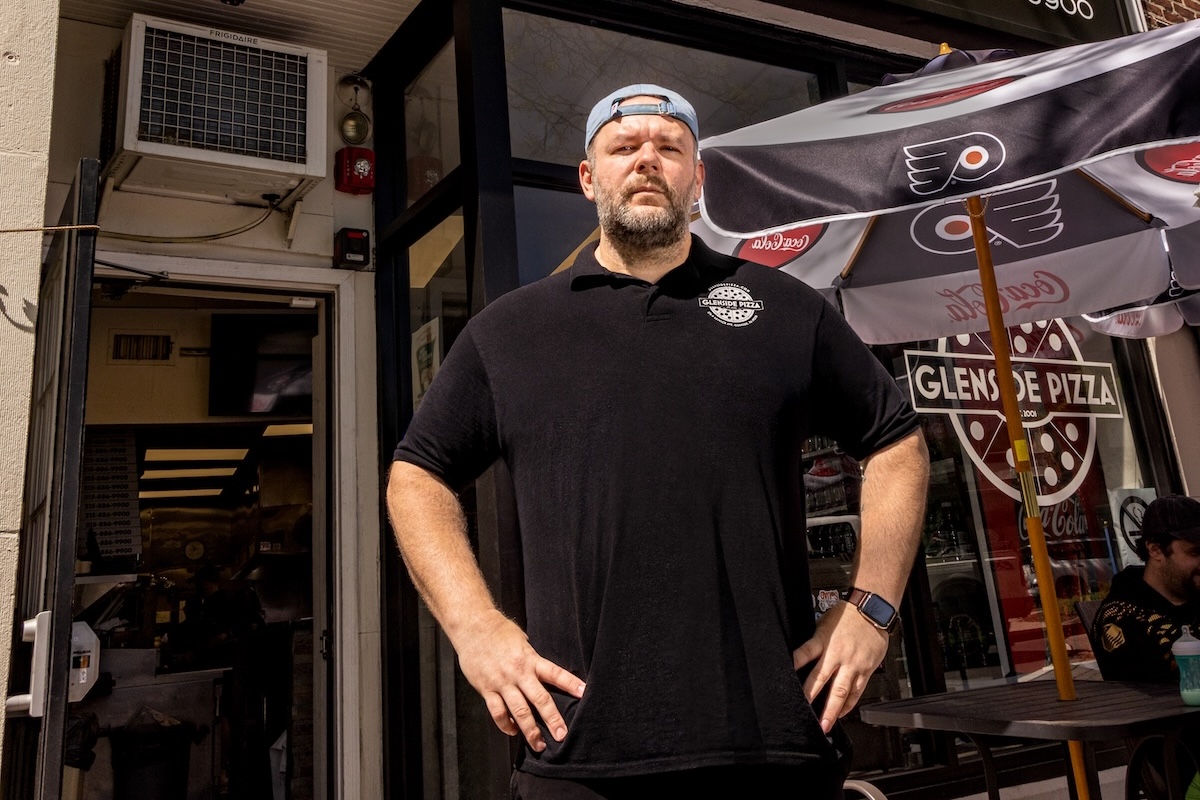 This photo shows Bill Zones, dressed in all black, standing in front of his pizza shop with his hands on his hips.