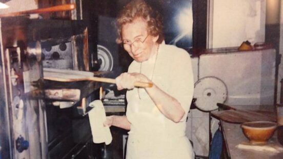 This washed out vintage photo shows Audrey Anderson looking at the camera as she peels a pizza into her oven.