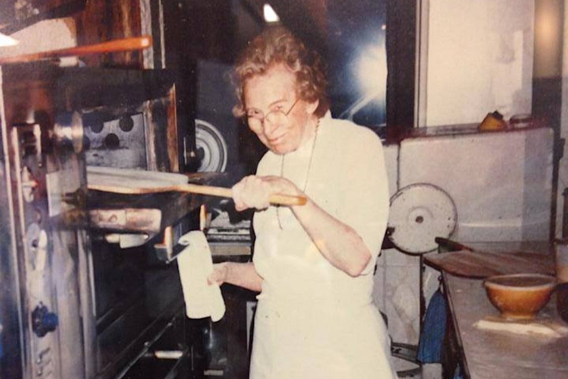 This washed out vintage photo shows Audrey Anderson looking at the camera as she peels a pizza into her oven.