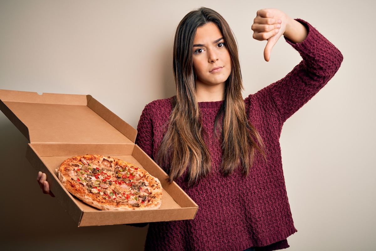 This photo shows a young woman holding a pizza delivery box and giving the thumbs-down sign.