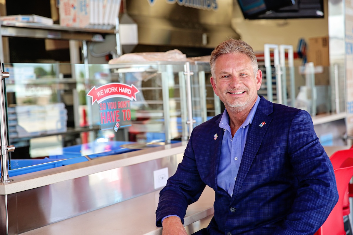 This photo shows Brian Bailey sitting inside his 100th Domino's store and smiling proudly.