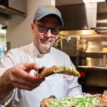 This photo shows Wylie Dufresne, wearing a white chef coat, holding up a slice of pizza for the camera.