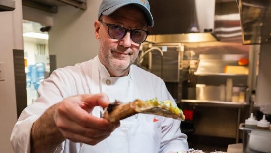 This photo shows Wylie Dufresne, wearing a white chef coat, holding up a slice of pizza for the camera.