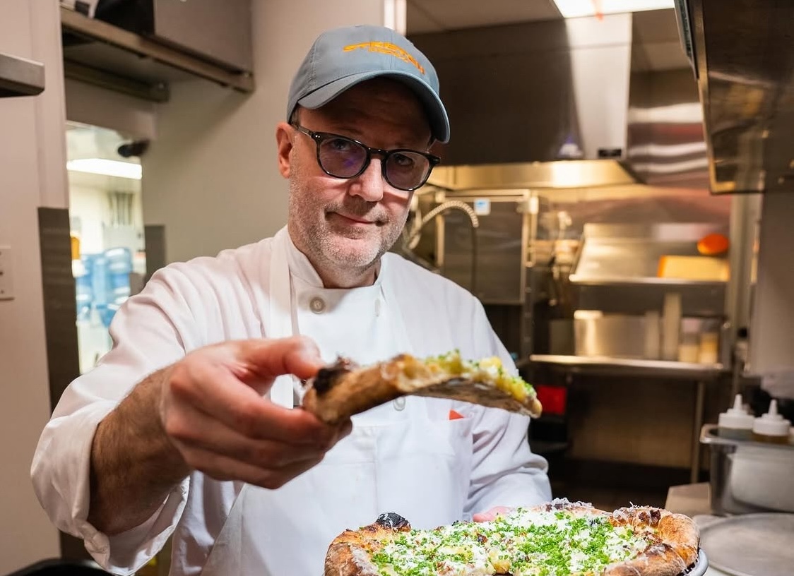 This photo shows Wylie Dufresne, wearing a white chef coat, holding up a slice of pizza for the camera.