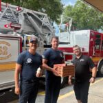 This photo shows three firemen, standing in front of a large fire truck, holding boxes of pizza from Jet's Pizza.