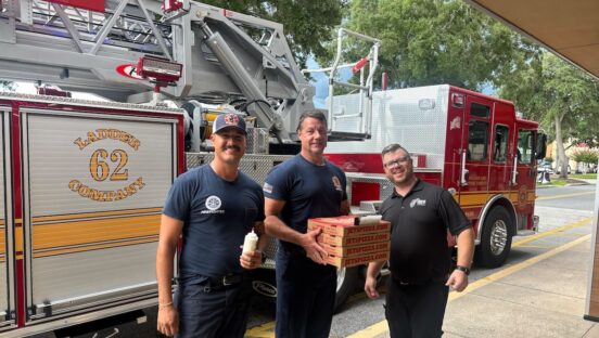 This photo shows three firemen, standing in front of a large fire truck, holding boxes of pizza from Jet's Pizza.