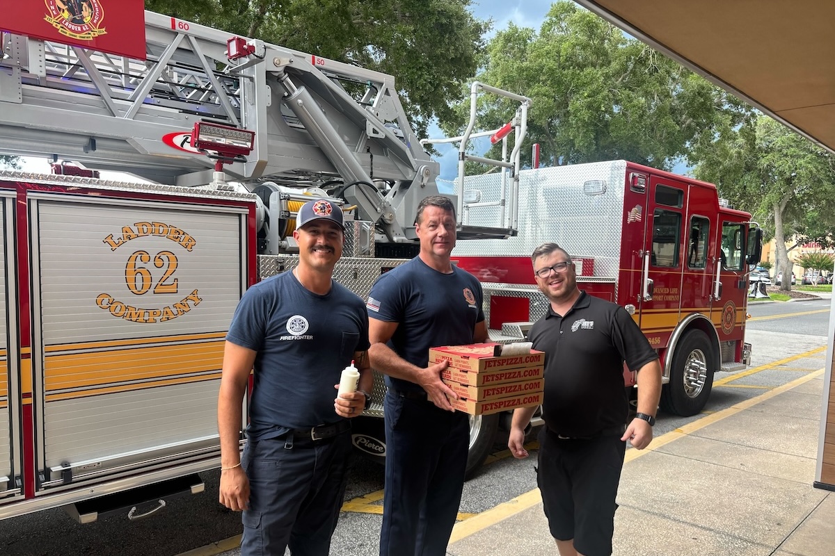 This photo shows three firemen, standing in front of a large fire truck, holding boxes of pizza from Jet's Pizza.