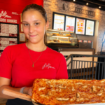This photo shows a young dark-haired woman in a red Ledo Pizza t-shirt holding up a pizza in the restaurant's dining room.