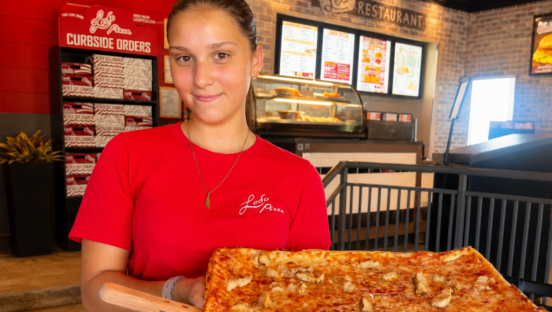 This photo shows a young dark-haired woman in a red Ledo Pizza t-shirt holding up a pizza in the restaurant's dining room.