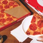 This photo shows a woman's hand holding a slice of Marco's New York-style pizza in a paper plate with the remainder of the whole pie in a box.