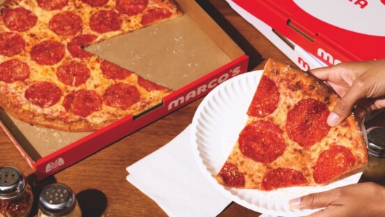 This photo shows a woman's hand holding a slice of Marco's New York-style pizza in a paper plate with the remainder of the whole pie in a box.