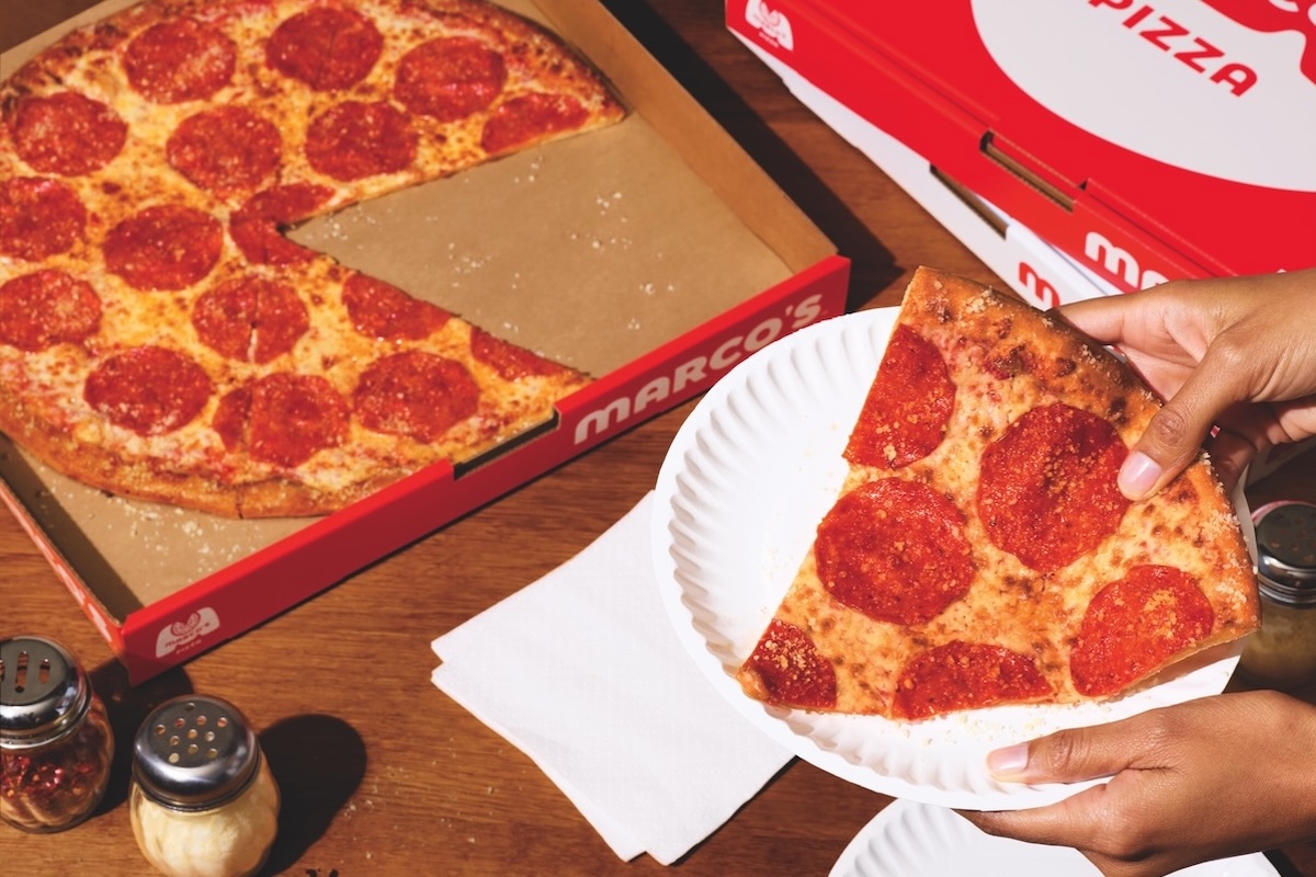 This photo shows a woman's hand holding a slice of Marco's New York-style pizza in a paper plate with the remainder of the whole pie in a box.
