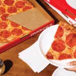 This photo shows a woman's hands holding a slice of New York-style pepperoni pizza in a paper plate.