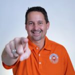This photo shows Matt Plapp in his trademark orange shirt smiling and pointing confidently at the camera.